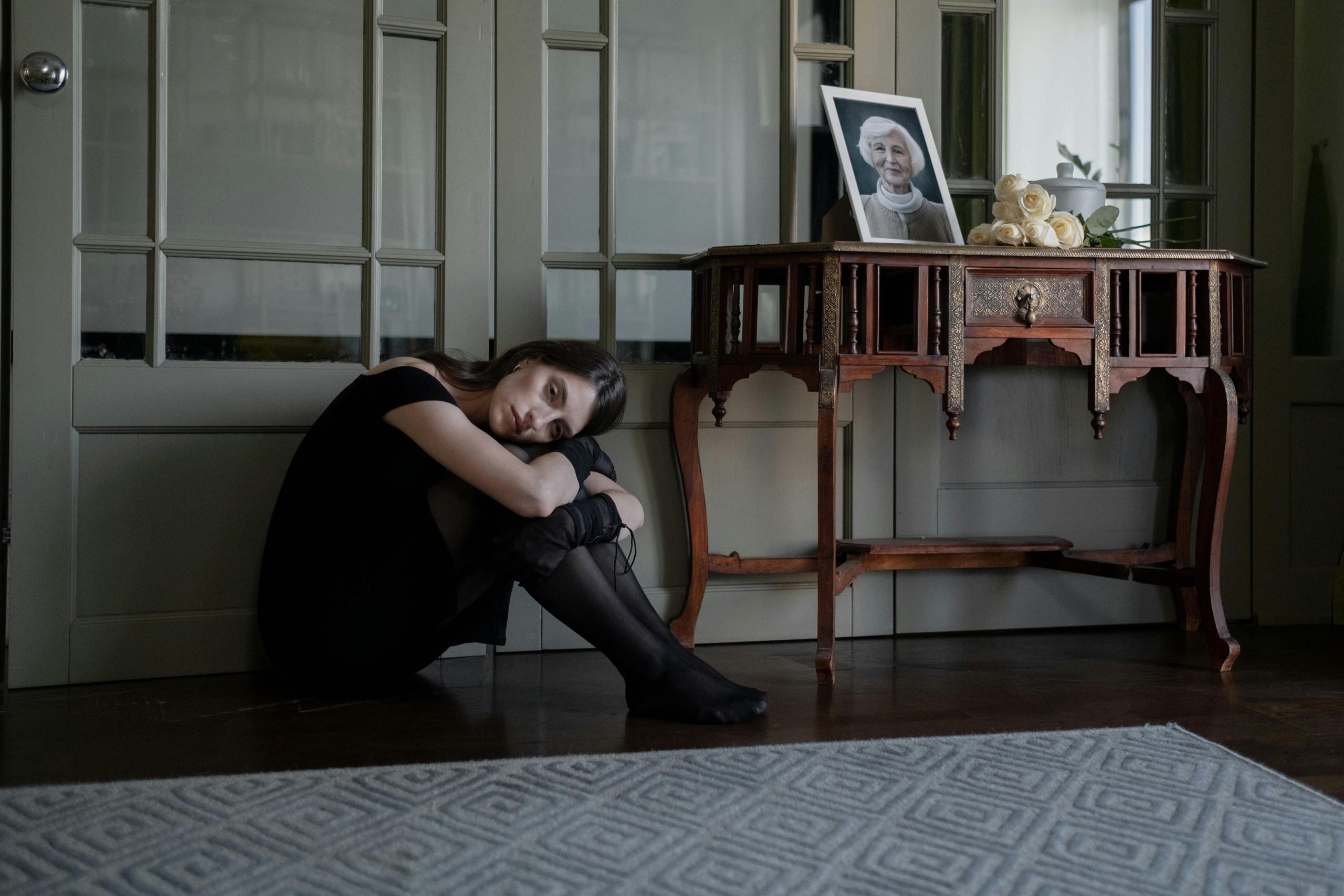 A solitary woman in black attire sits on the floor in mourning inside her home.