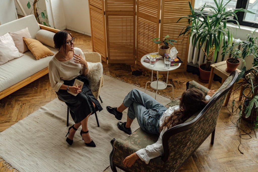 woman-in-white-long-sleeve-shirt-sitting-on-brown-wooden-armchair-5217852 Two women engaged in a psychotherapy session in a warm, inviting interior with plants and natural lighting.